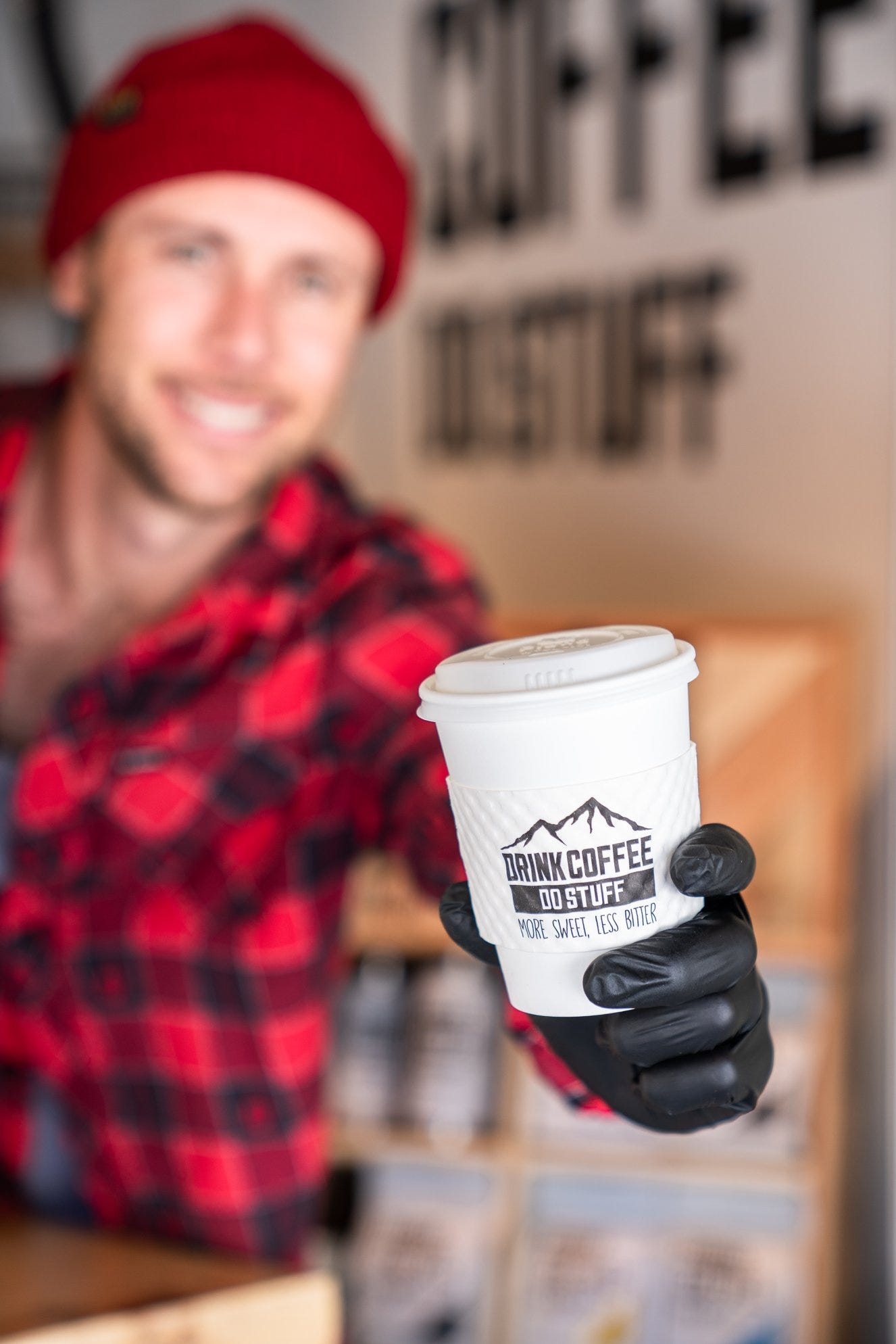 A man in a red buffalo check flannel is blurred in the background holding a white takeaway coffee cup 