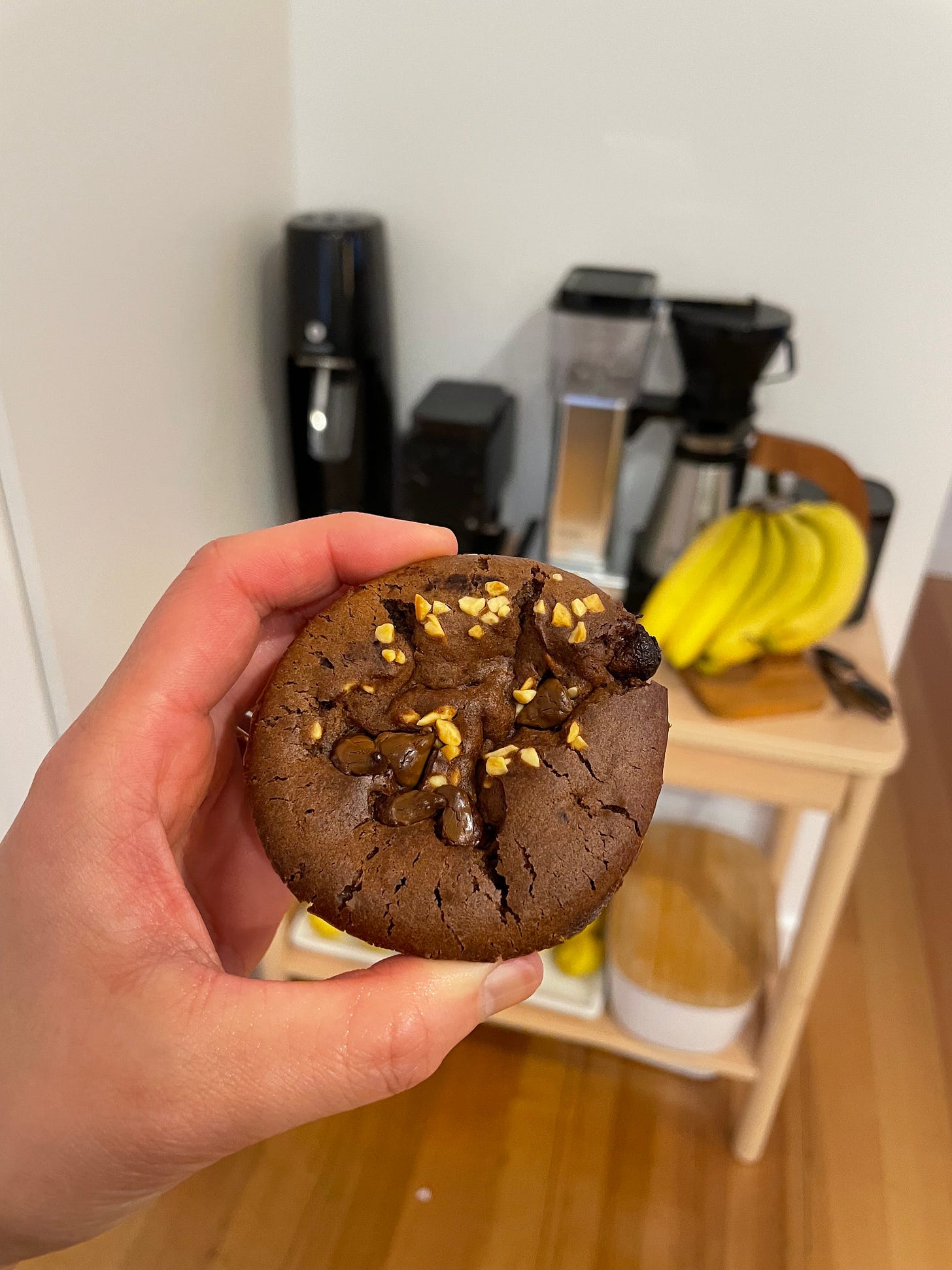 Chocolate muffin with chopped peanuts and choc chips sprinkled on top. A coffee machine and bananas on a kitchen bench in the background.