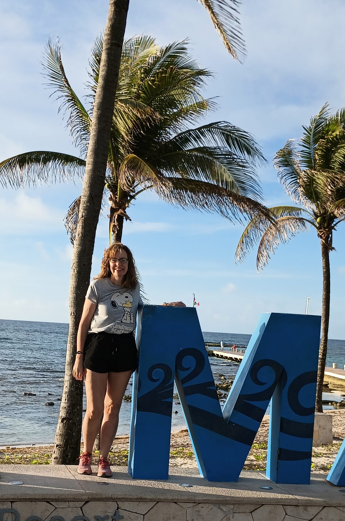 woman with light skin, dark hair, and glasses standing i fromt of beach with palm trees and next to large blue letter M.