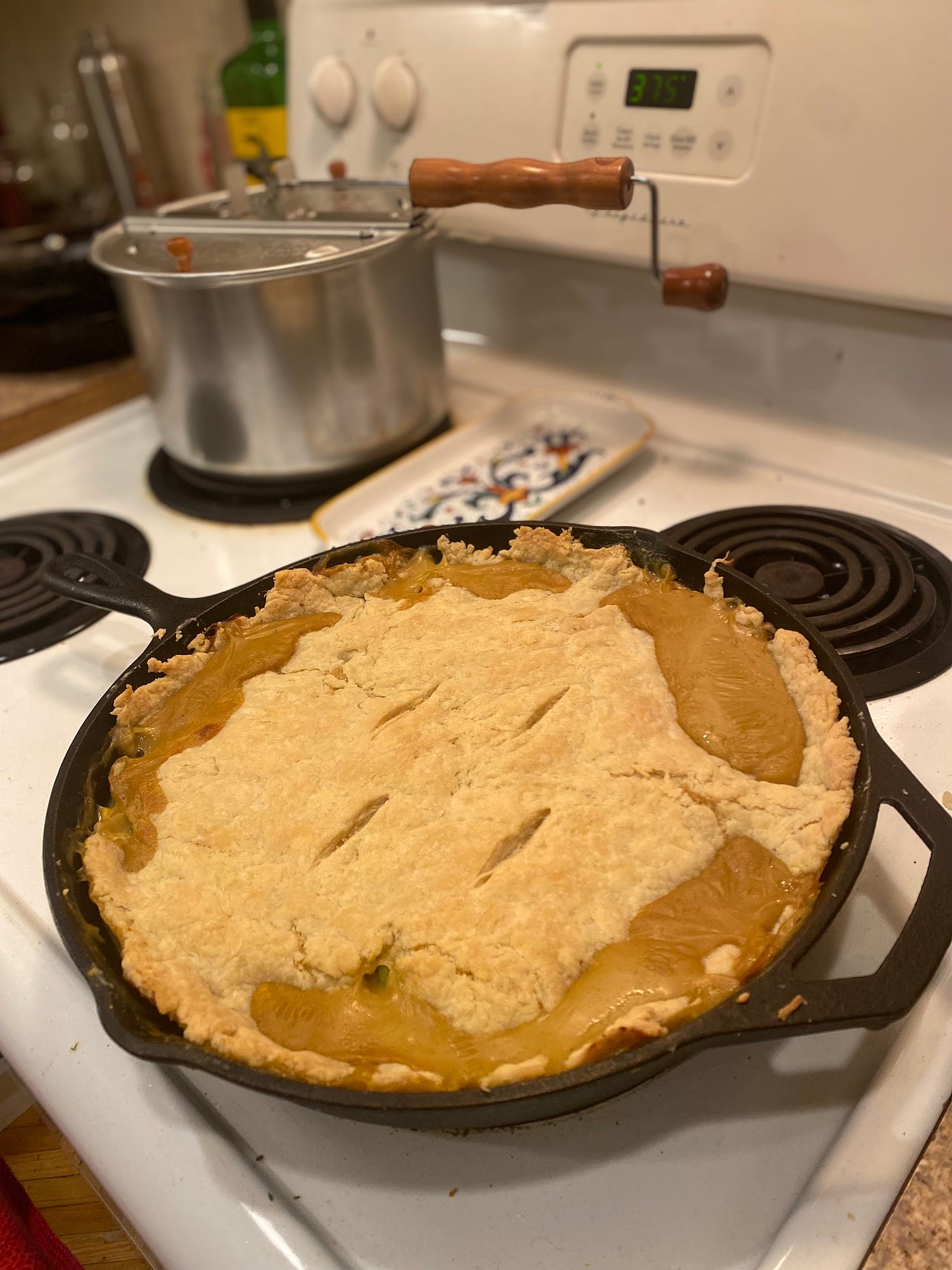 On the stovetop, a large cast-iron pan of the pot pie described above, the crust with a few scores in the centre and broken in a few places near the edges. Filling has also leaked up over the top.
