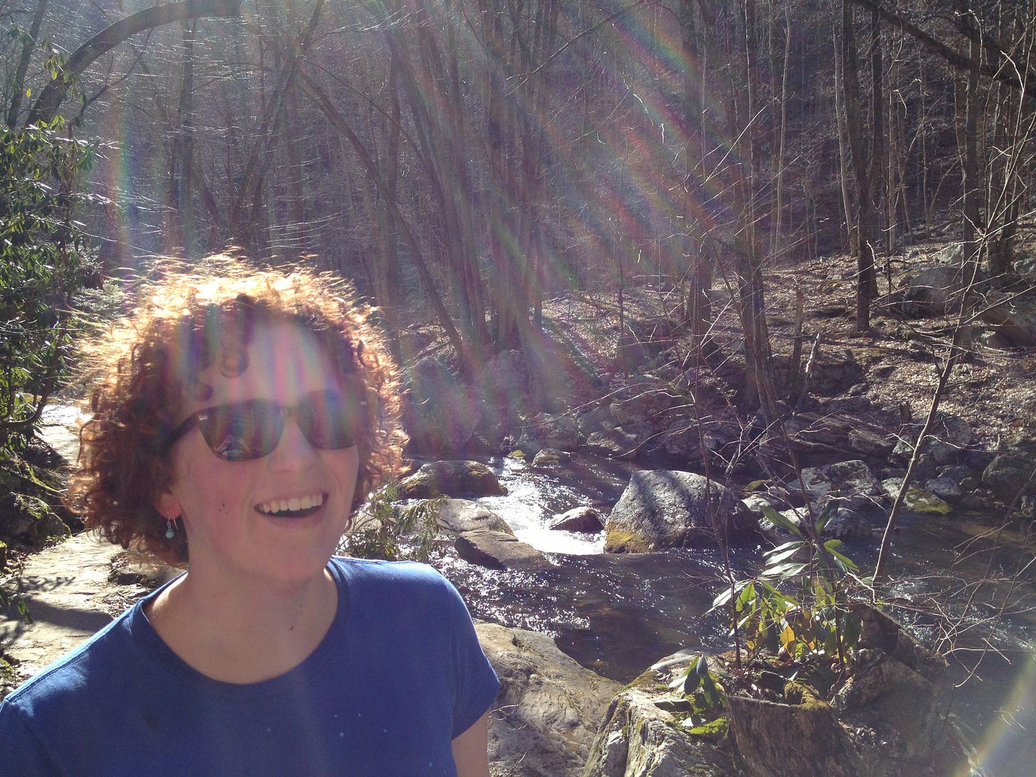 Joan wears sunglasses and a big smile. The sun and a rainbow glow across her. They stand in a forest, a creek runs beside them.