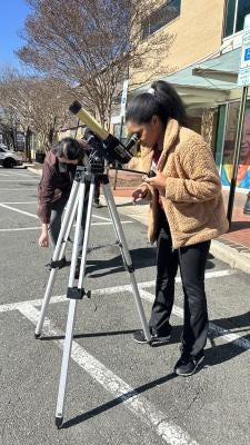 "An adult wearing a beige coat looking through a telescope outside.