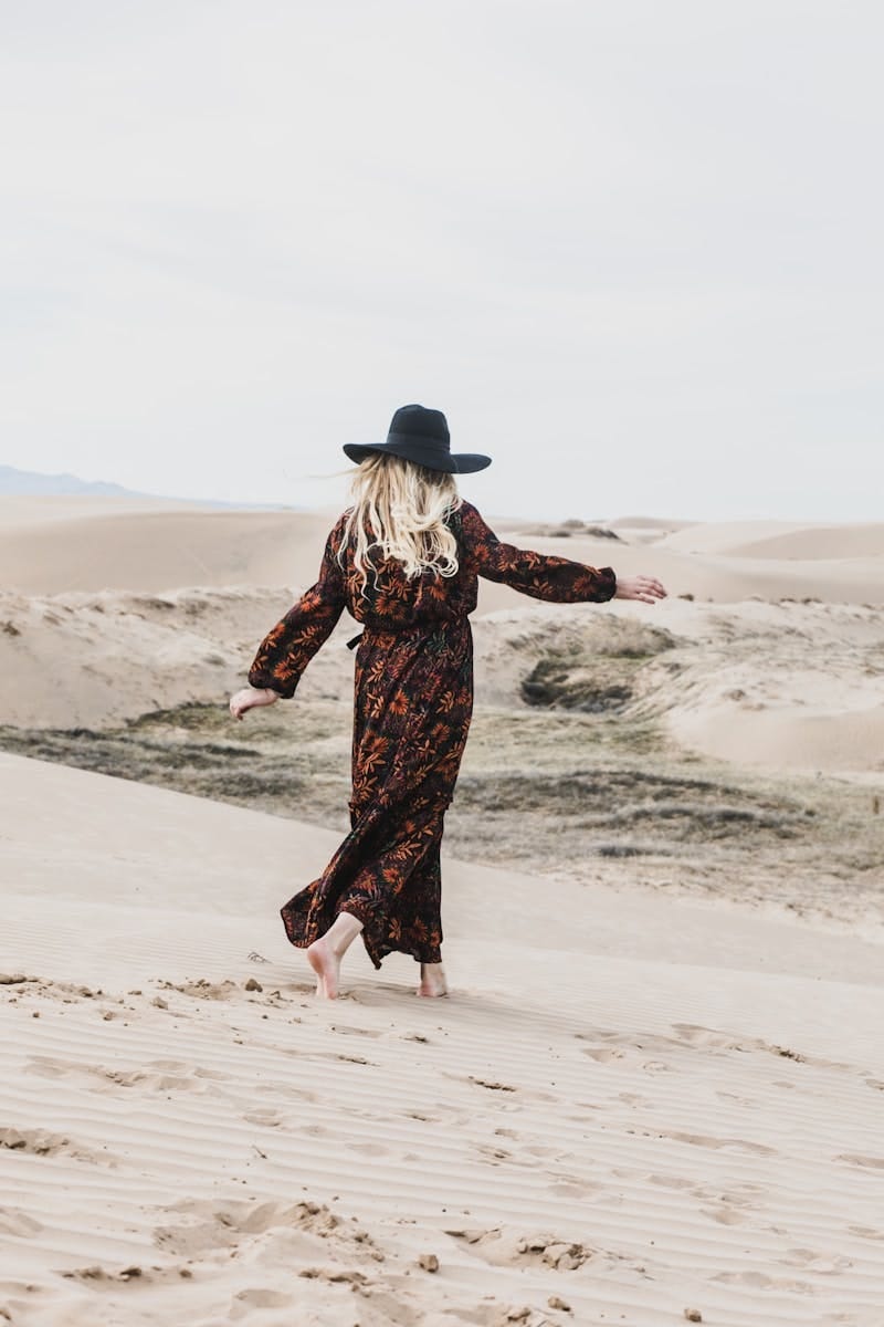 Photo of the back of a woman with blond hair, wearing a long dress and black hat, walking barefoot on the beach
