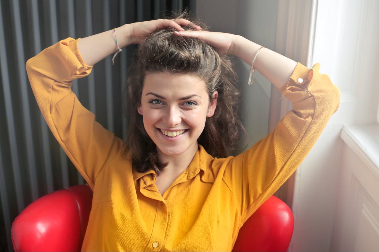 A young woman in a mustard-colored button shit has her hands on her head and a big smile.