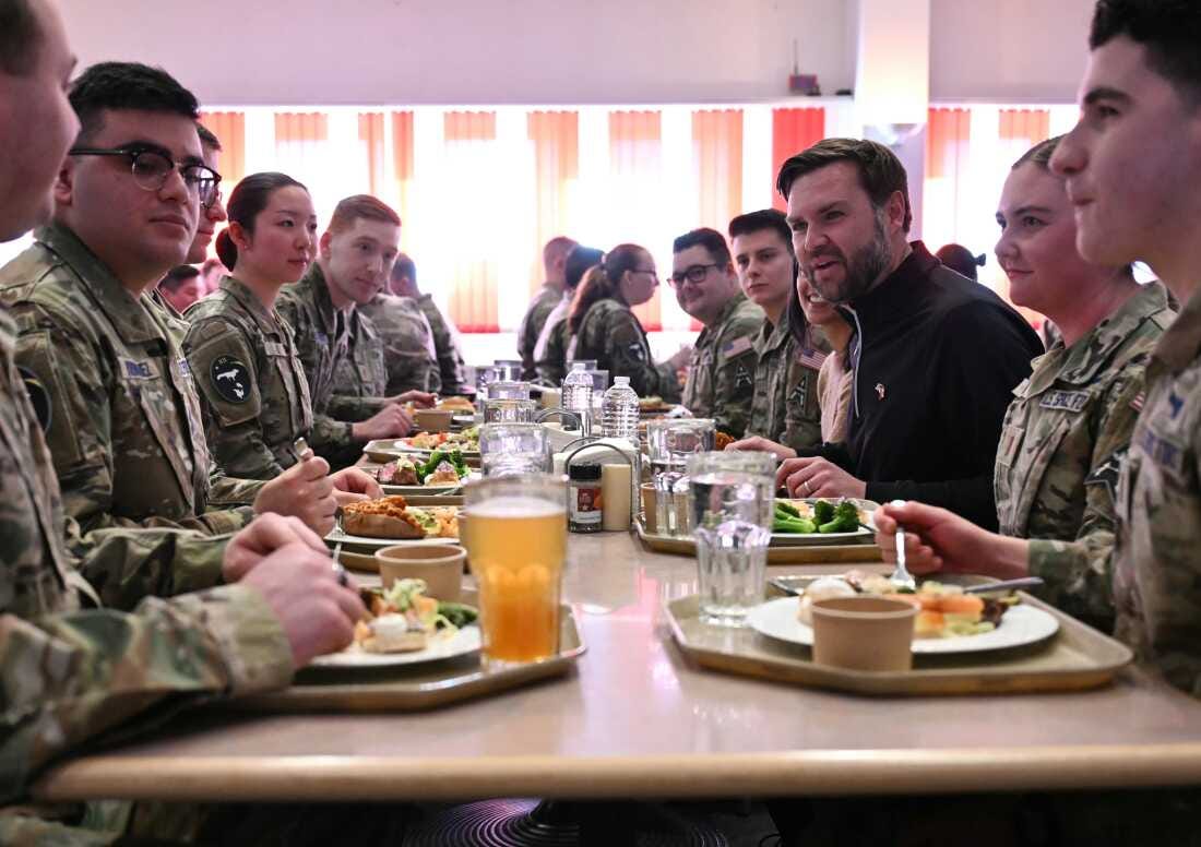 In this photo, Vice President Vance is seated on the right side of a long table. His wife is seated next to him. U.S. service members wearing uniforms sit at the table's other seats. They all have food and drinks on cafeteria trays in front of them.