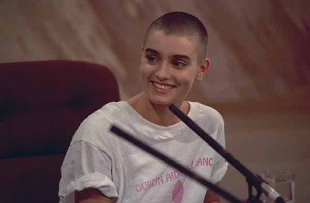 Sinéad O’Connor, immagine a colori dalla TV irlandese. Sorride. Indossa una maglietta con la scritta “Dublin AIDS Alliance”