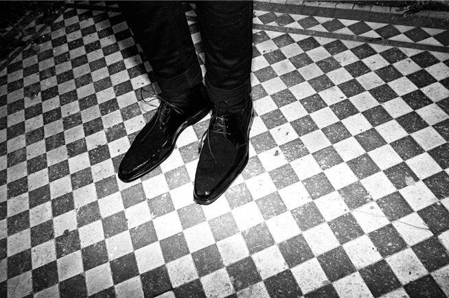 Black and white photo of a man, showing the knees down, with slim cut trousers and a pair of Derby shoes with suede vamp and stylishly pointed toe.