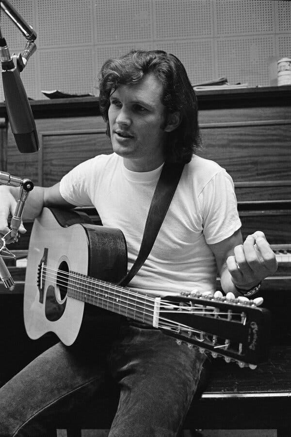 A black-and-white photo of Kris Kristofferson, in a white T-shirt, sitting on a piano bench faced away from the instrument, an acoustic guitar on his lap and a microphone poised in front of his face.