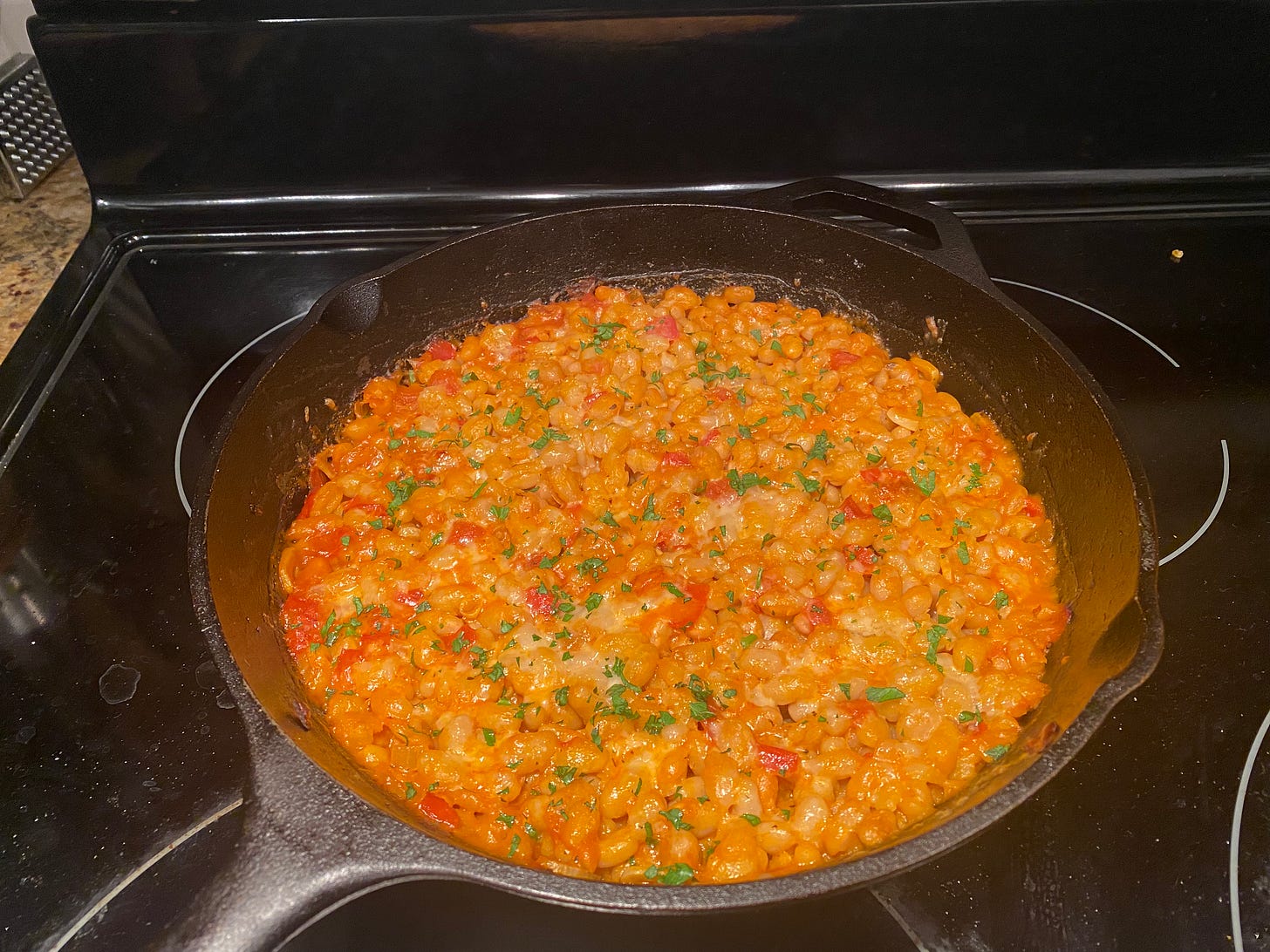 A cast iron pan filled with the bean dish described above. It's navy beans with pieces of tomato throughout, spots of lightly browned cheese, and sprinkles of parsley.