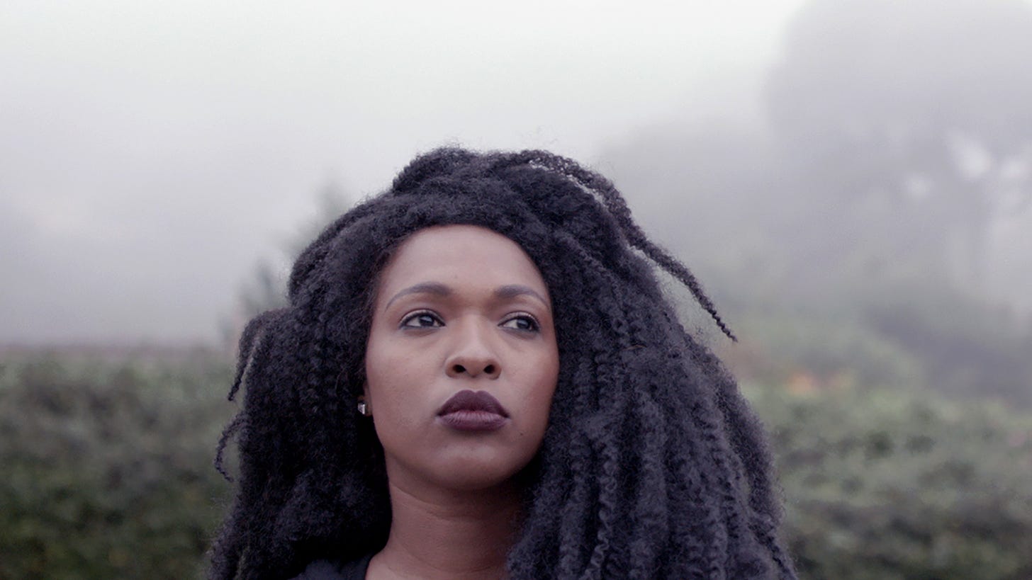 A Black woman looks out past the camera against a foggy hillside.