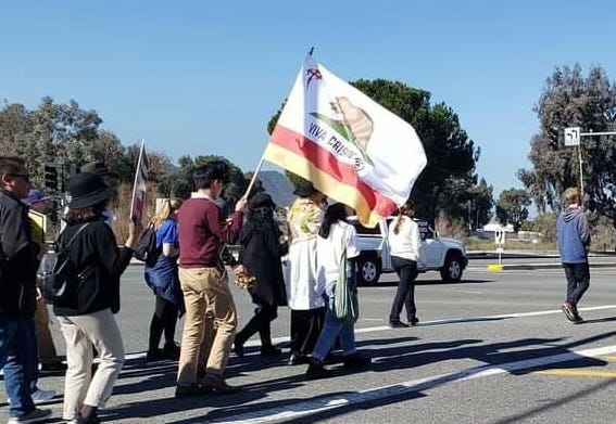 People walking along the street in a walk for life. Crucifix and California flag that reads viva Cristo Rey