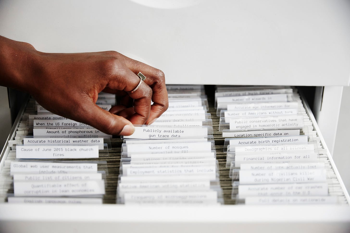 The Library of Missing Datasets by Mimi Onuoha showing a light grey maxi-filing cabinet filled with labelled but empty files. The hand of a Black person is show extracting one file labelled “Publicly available gun trace data”.