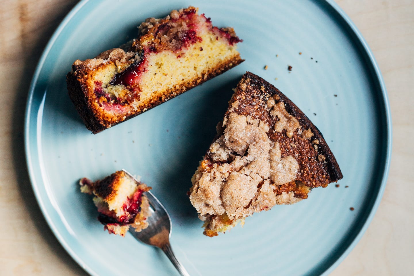 Two slices of plum cake on a plate, one is tipped on the side to show the crumb and jammy filling, and the other is seen from overhead with a bite arranged on a fork alongside. 