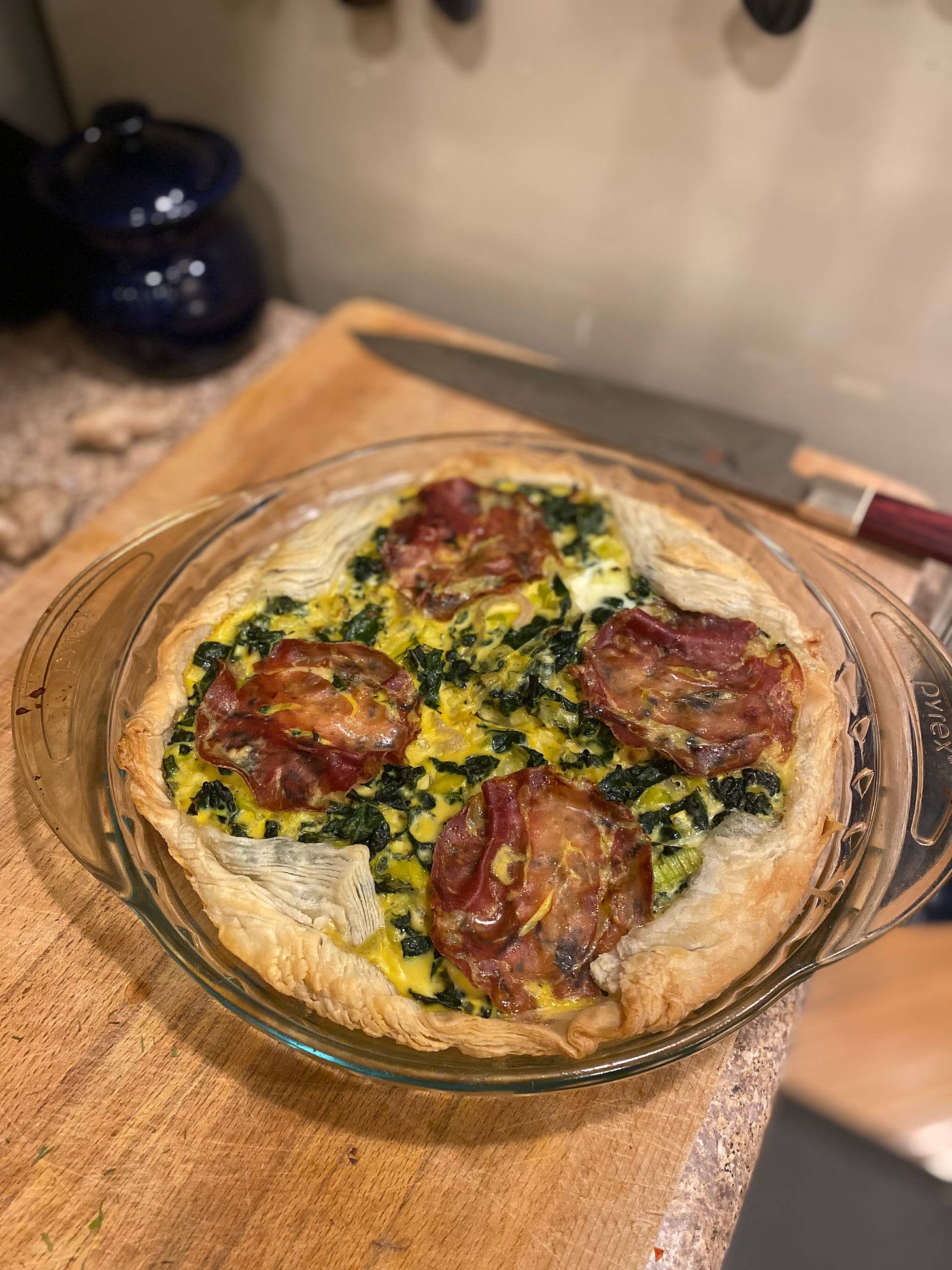 A puff pastry quiche with leeks and dark green kale throughout. On top are four lightly crisped slices of prosciutto-salami. It's in a glass pie pan resting on a cutting board next to a knife.