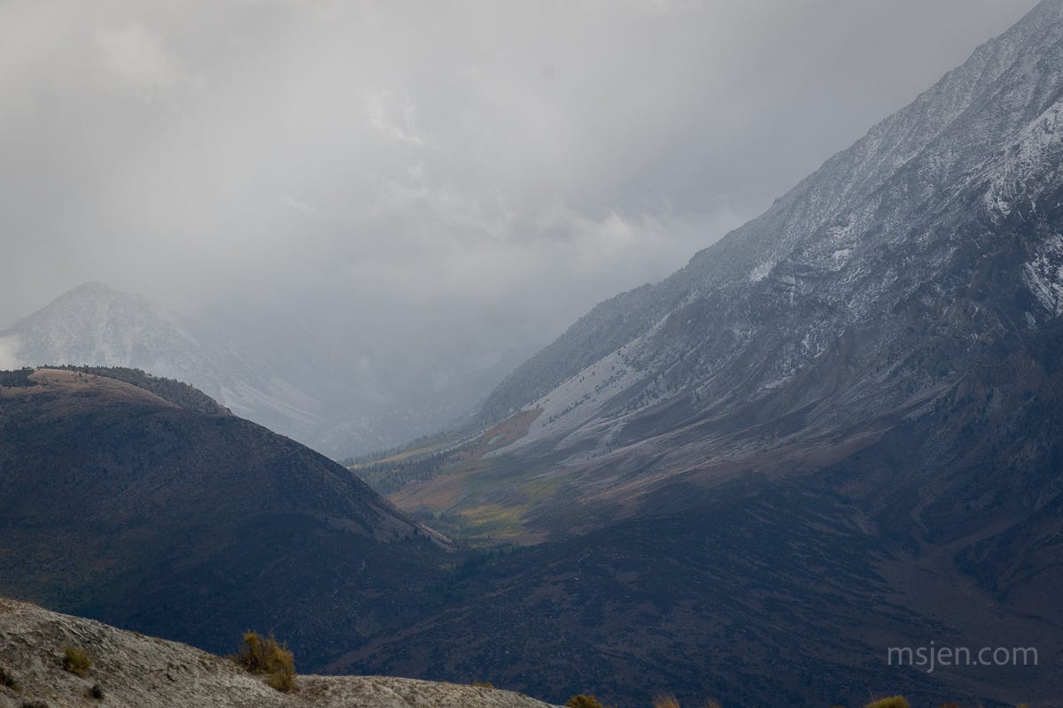 Snow falling in the high mountains as a wee bit of sunlight breaks through the clouds to illuminate yellow & orange fall aspen trees in a high up valley in the mountains.