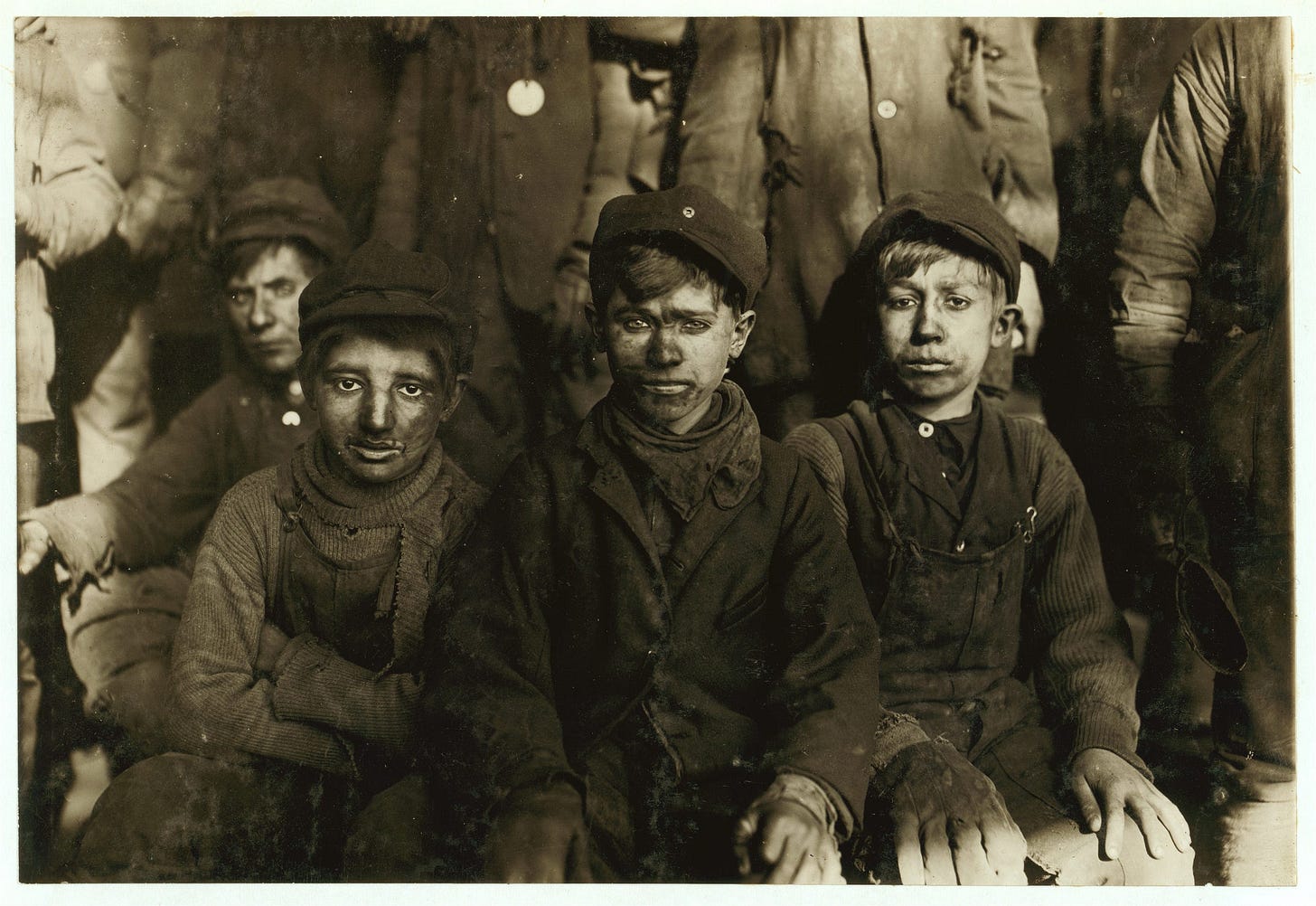 Lewis Hine b/w Library of Congress photo from 1911 showing child laborers, several young, seated  ‘breaker boys.’ The photo depicts small boys, 8–10 years old, in dirty clothing, faces sneared with coal dust, working in an anthracite coal mine in eastern Pennsylvania.