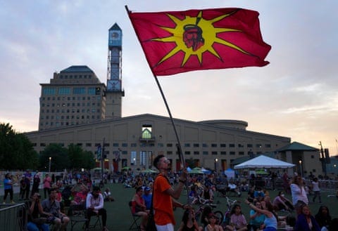 Man in sunglasses and orange shirt waves the Six Nations flag, a red flag with a yellow sun and illustration of a chief in the center, at an outdoor gathering