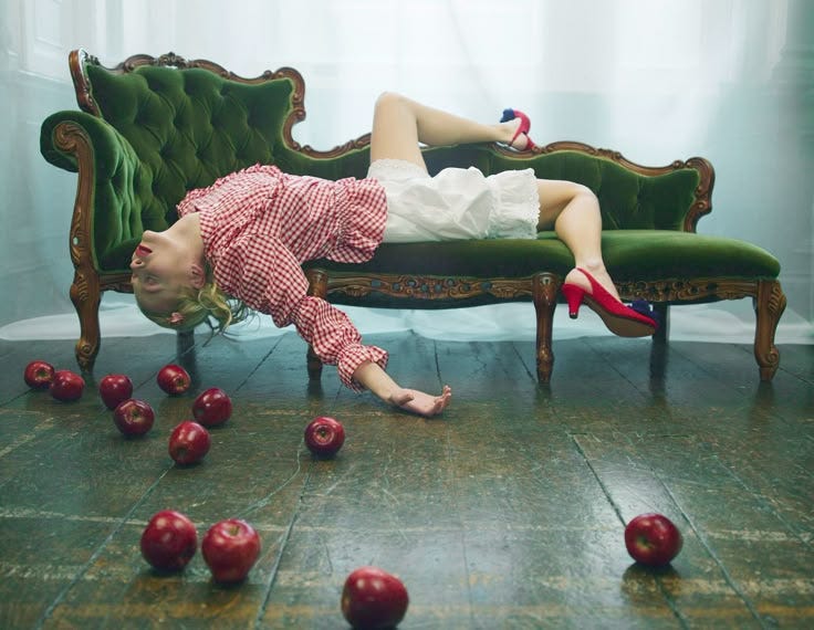 A woman in red heels and a gingham shirt lies dramatically across a vintage green velvet sofa, surrounded by scattered red apples on a wooden floor.