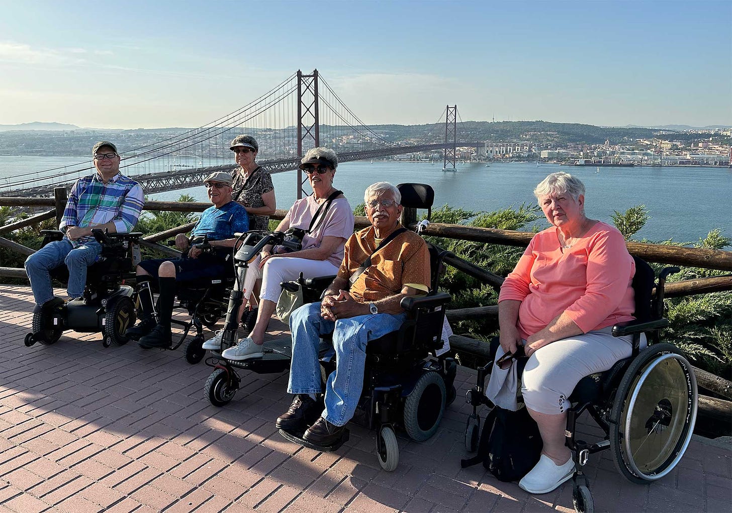 Group of Wheelchair Travel readers with John in front of a bridge styled like the Golden Gate overlooking a large river and the city of Lisbon.