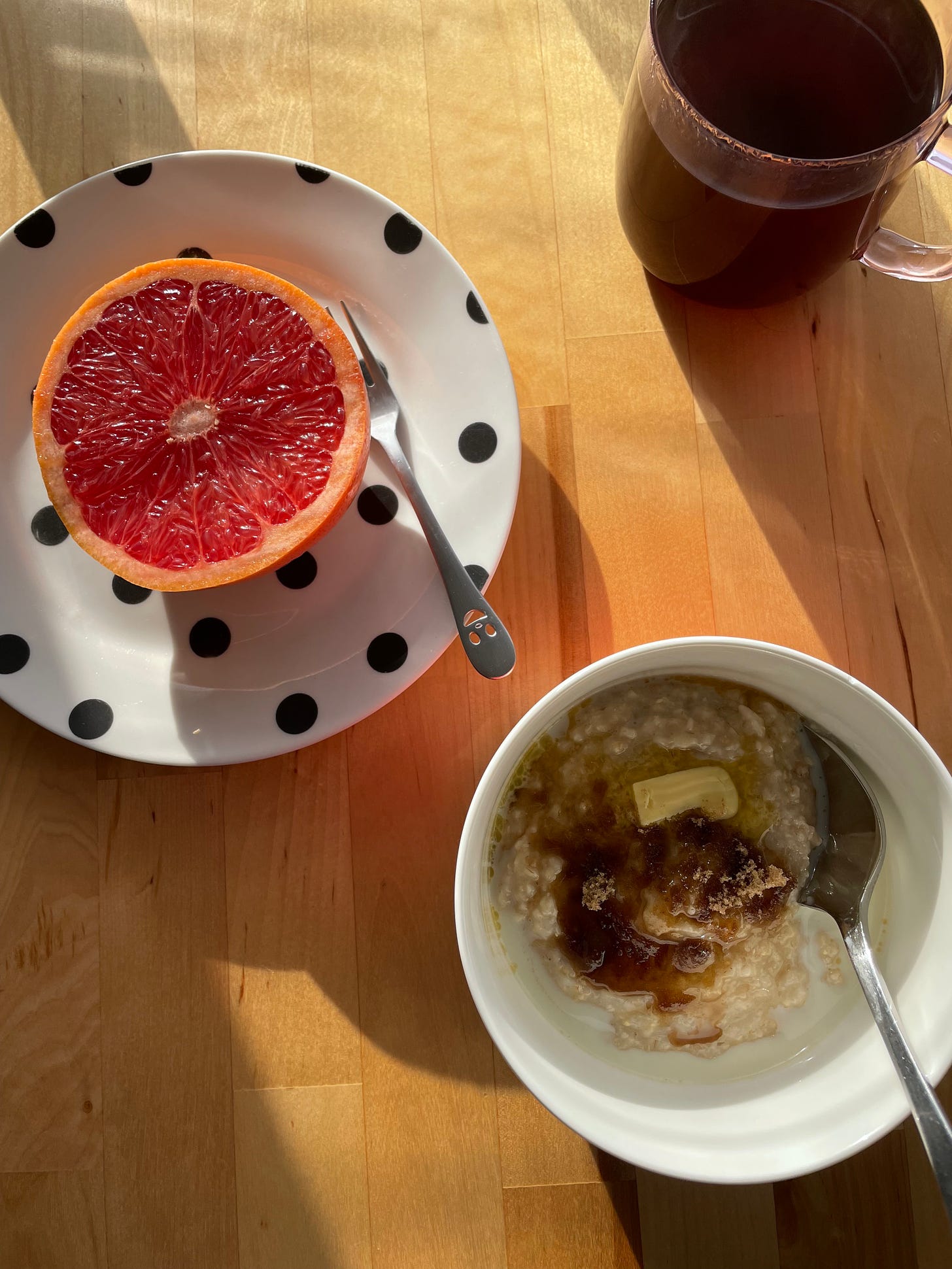 Breakfast table with a bowl of oats topped with butter and brown sugar. A red grapefruit half sits on a spotty plate nearby, with a cup of filter coffee in a translucent pink mug.