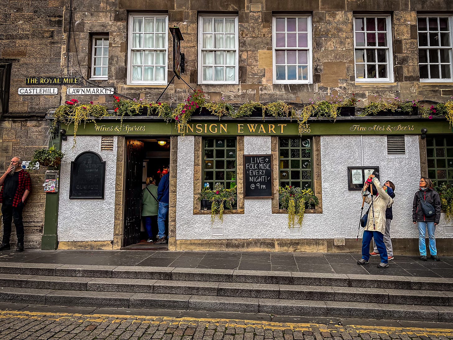 Exterior view of the pub, green sign with yellow lettering, with flowers from hanging baskets around the entrance. Tourists outside taking photos of the castle, which is out of shot.