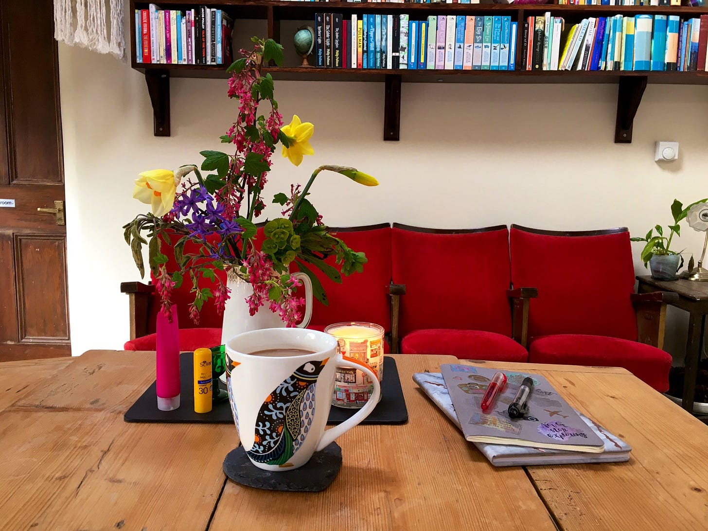 Kitchen table, with notebooks, pens, coffee and a vase of flowers. In the background, theatre seats and the bottom of a set of wall-mounted bookshelves.