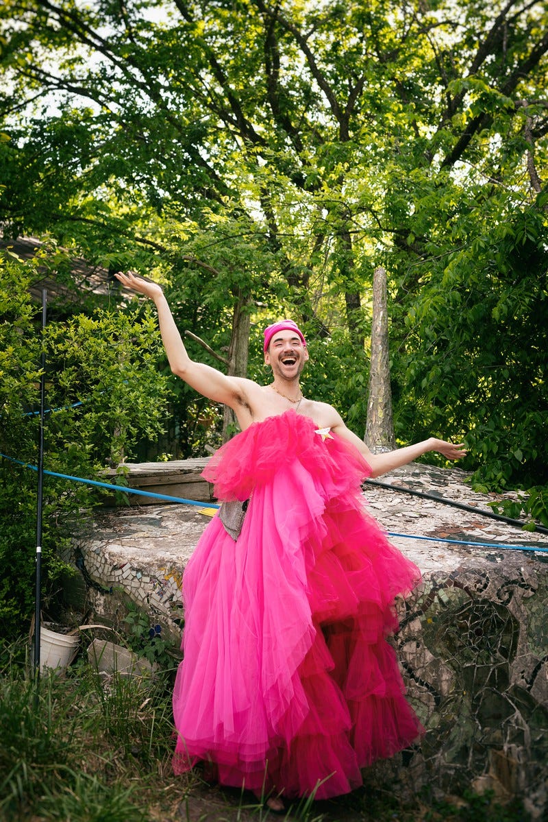 a photograph of a lush green canopy of woods. in the middle is me, a scruffy white person with a big wide open-mouthed smile and arms outstretched to either side gesturing to an entire surrounding. i’m wearing layers of pink tulle ruffles, so much fabric that my purse is swallowed up on one side, and a pink knit beanie hat. behind me there is a mosaic-covered water cistern built into the side of a hill and a few cables or tubes running across it.