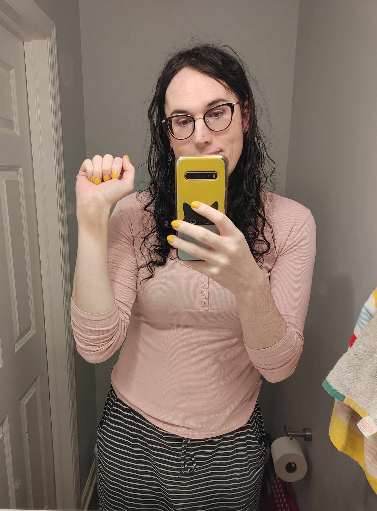 A mirror selfie of me, a pale white woman with wavy, chest-length black hair and horned glasses, holding up my right arm to show my bare wrist with a good 3-inch long bruise