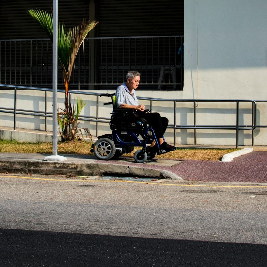 A man in a power wheelchair navigates toward a curb-cut in the sidewalk.