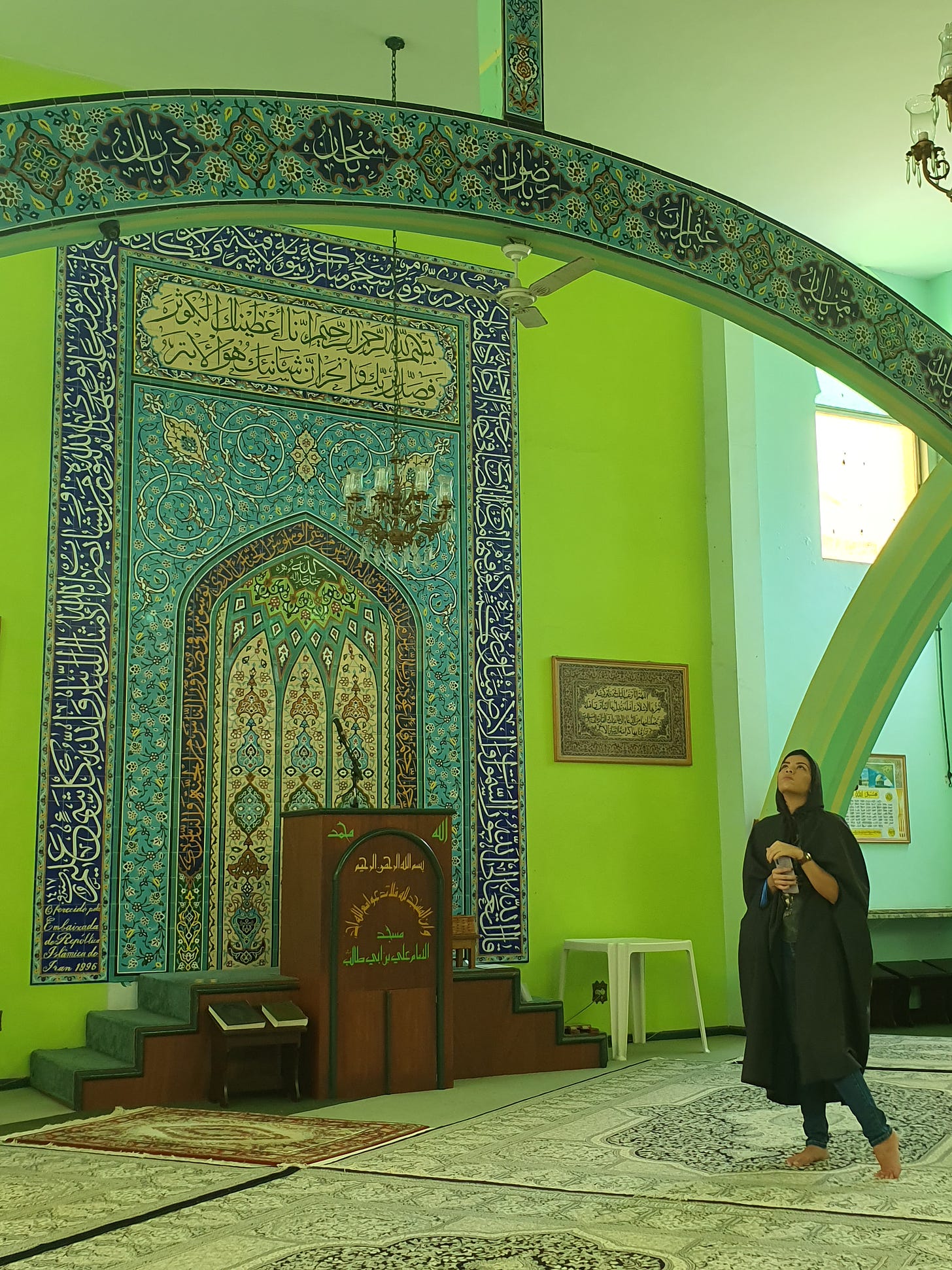 A young woman in dark abaya gazes upwards in front of the mimbar in a mosque with lime green walls and elaborate decor with Arabic script in blue, turquoise, cream, and gold. Small chandeliers hang from the high ceiling. Sunlight gently suffuses the room so that the colors glow. 