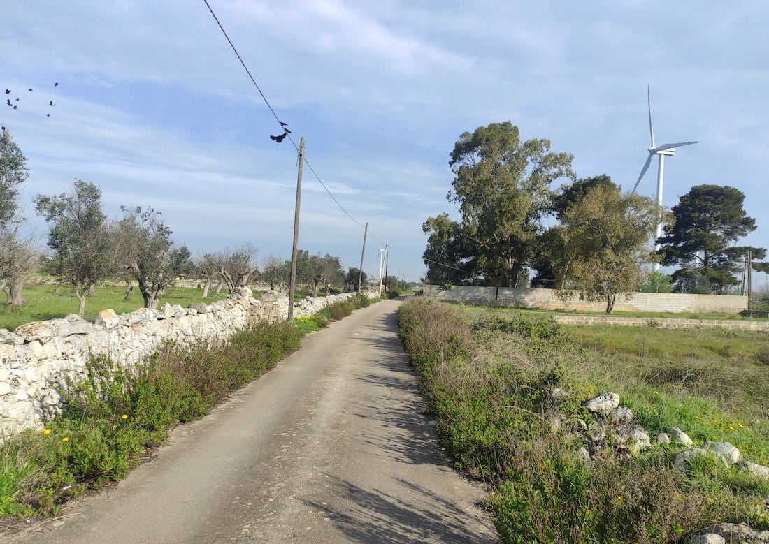 A narrow road bordered by a stone wall and grass, with an electricity generating windmill in the field beyond