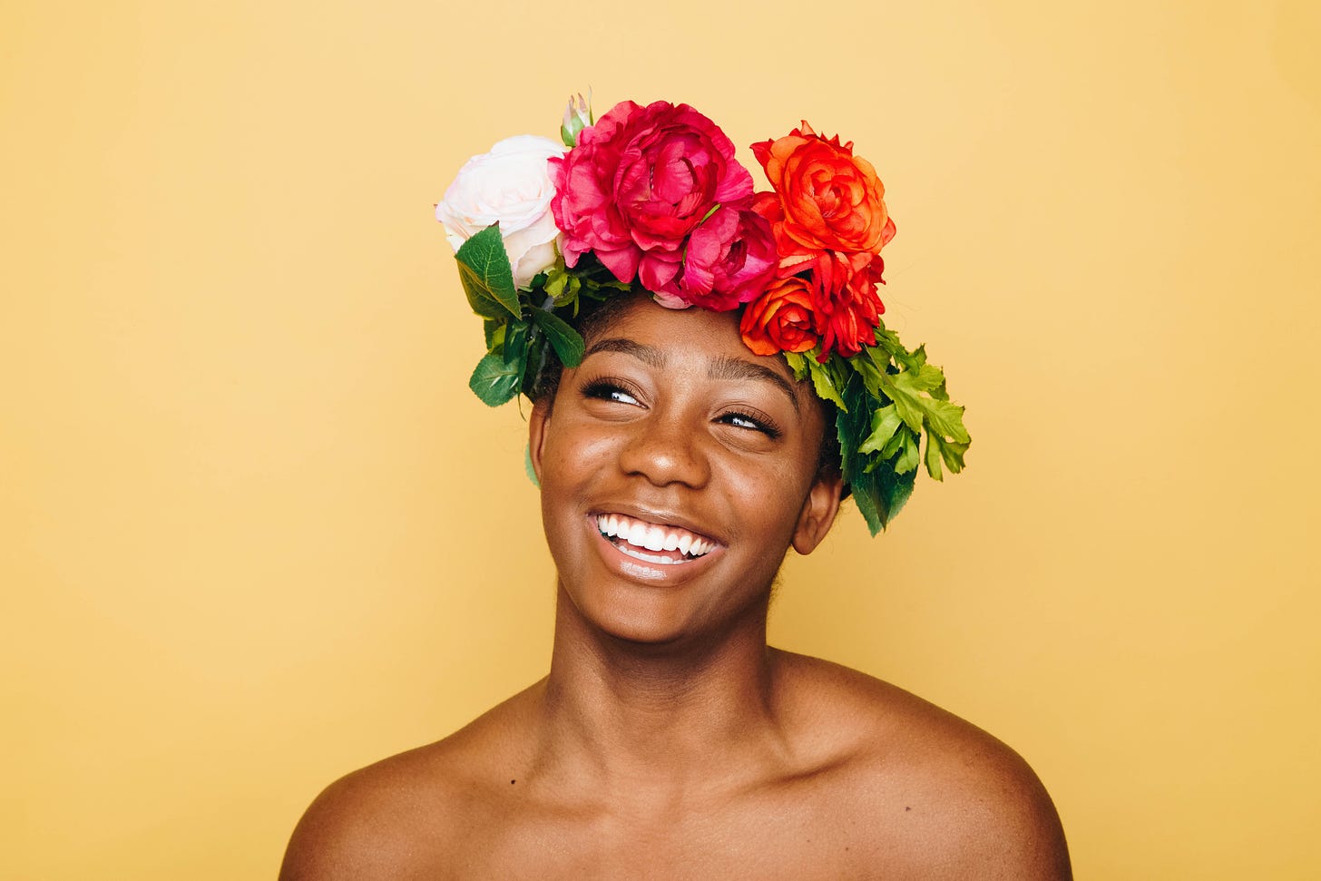 brown-skinned woman with a big smile, looking up and left, and a crown of flowers and yellow background