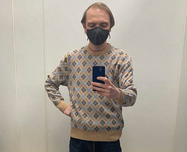 A white man in a mask stands in a fitting room wearing a tan argyle print sweater. He photographs himself with his phone.