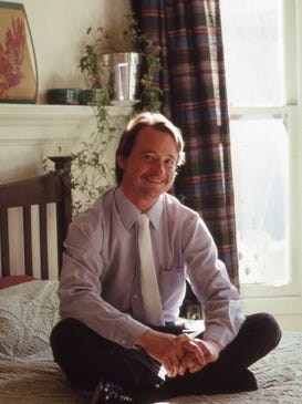 Lou Sullivan sitting cross-legged on a bed, in a shirt and tie, smiling.
