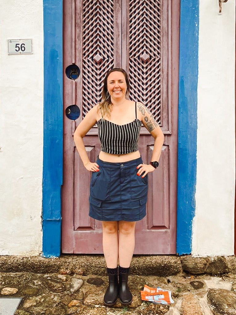 Uma mulher branca, com mãos na cintura, posta-se em frente a uma porta antiga. Veste galochas plásticas, uma saia e um cropped.