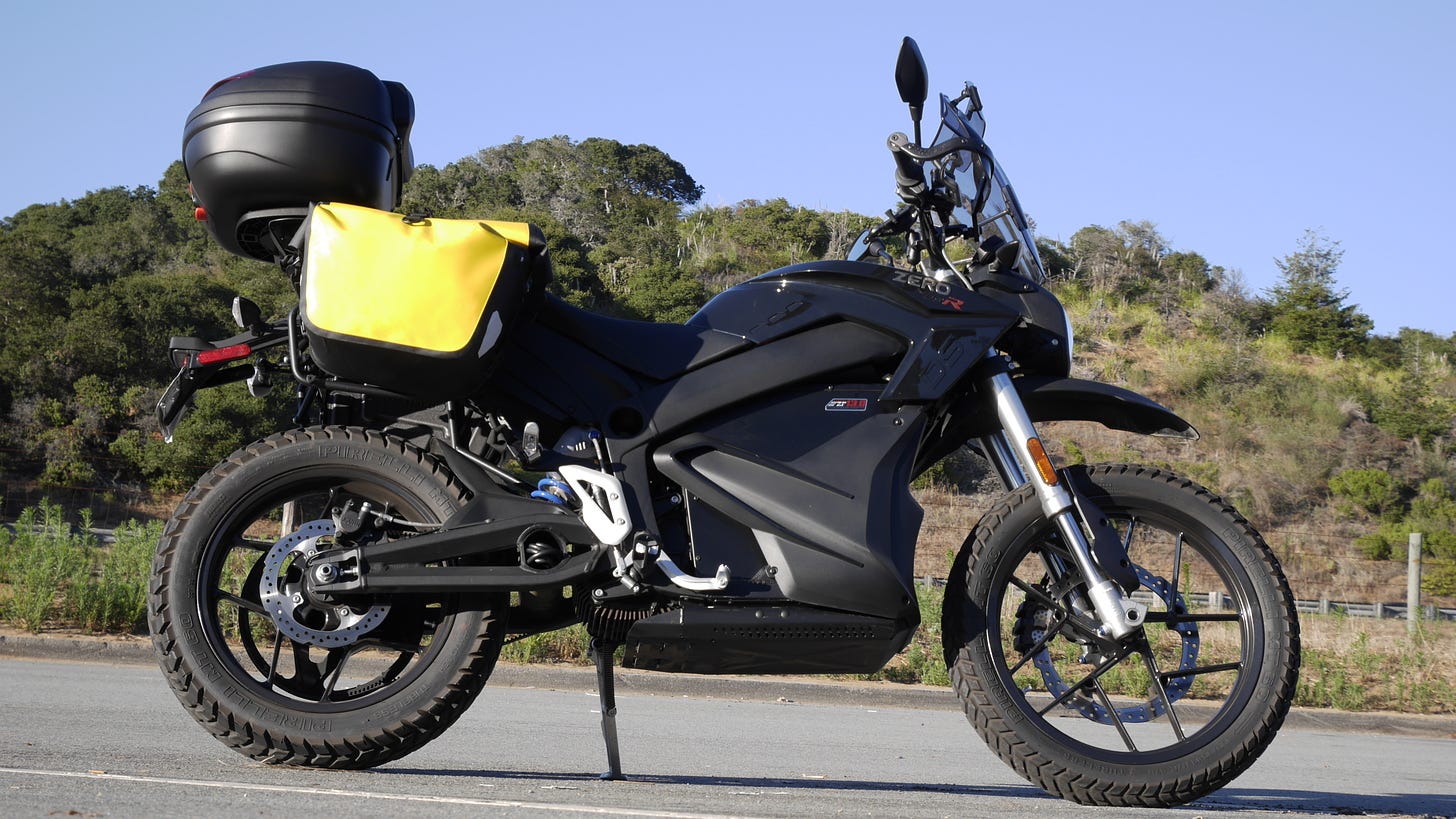 Side view photograph of a black electric motorcycle. with a large black topcase on the back and yellow saddlebags. The motorcycle has a windscreen and handguards. The battery of the motorcycle resides where the engine of a gasoline-powered motorcycle usually resides.