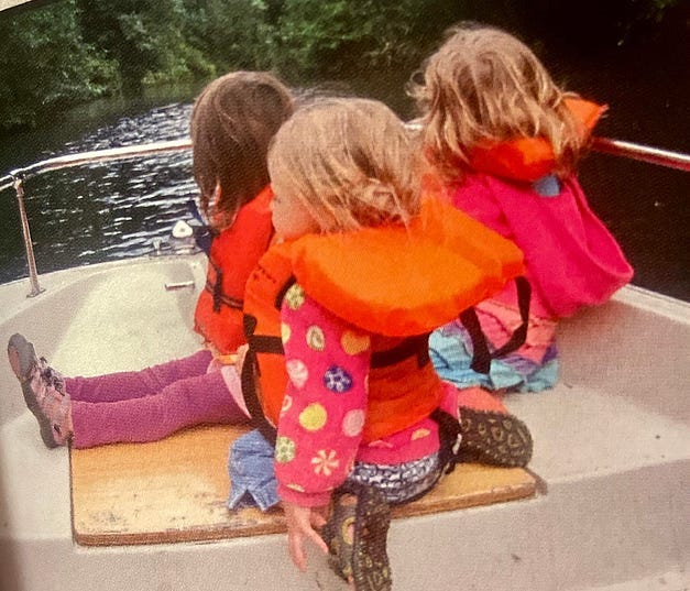 Three little girls, shot from behind, in orange life vests on a boat.