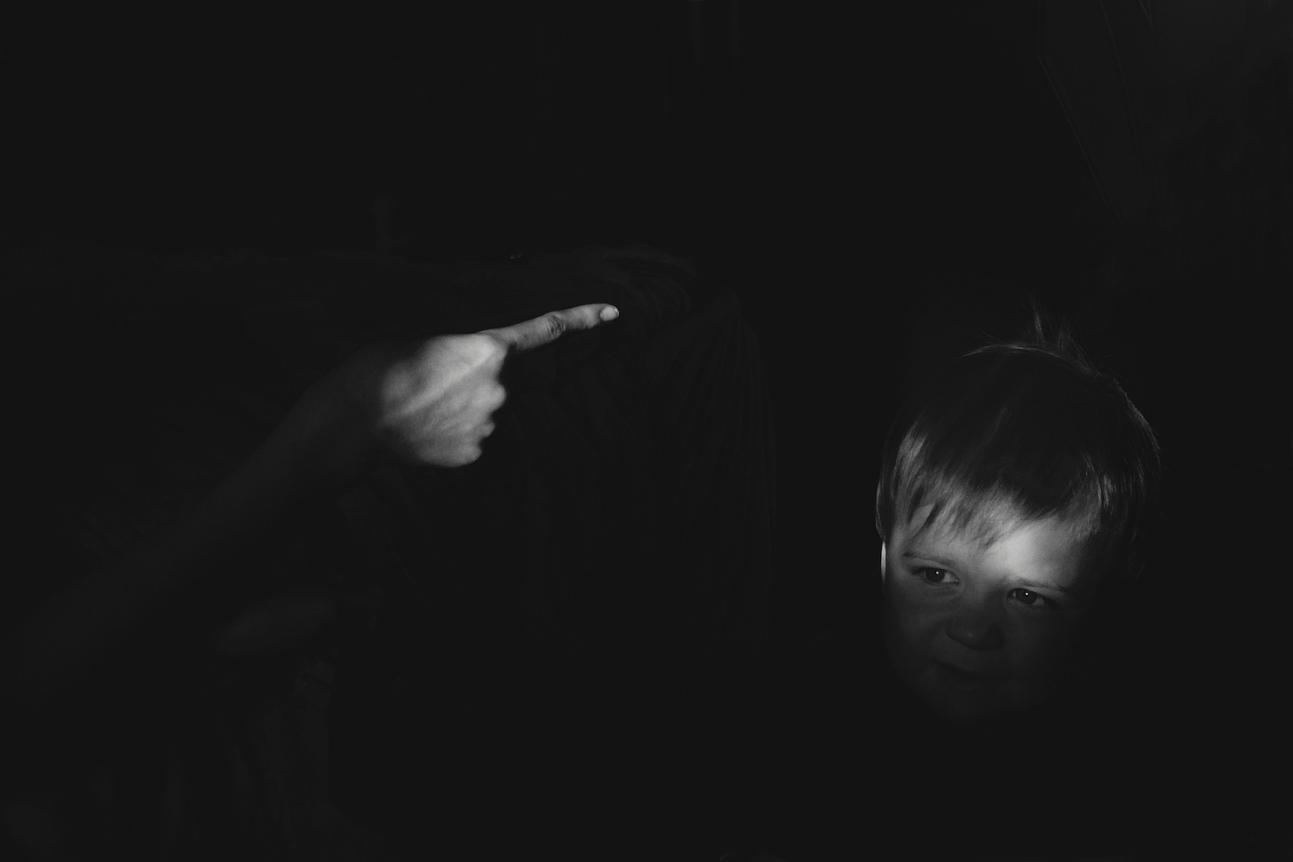 Black and white photo. On the left, the pointed finger of an adult is aimed toward a small, cowering, blonde child with fearful eyes.