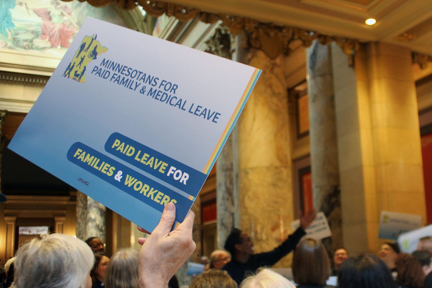 A hand holds up a blue sign with the Minnesota state outline reading "Minnesotans for paid family & medical leave" and "paid leave for families & workers" behind the sign is a crowd of people, one person is raising their hand toward the right side of the image