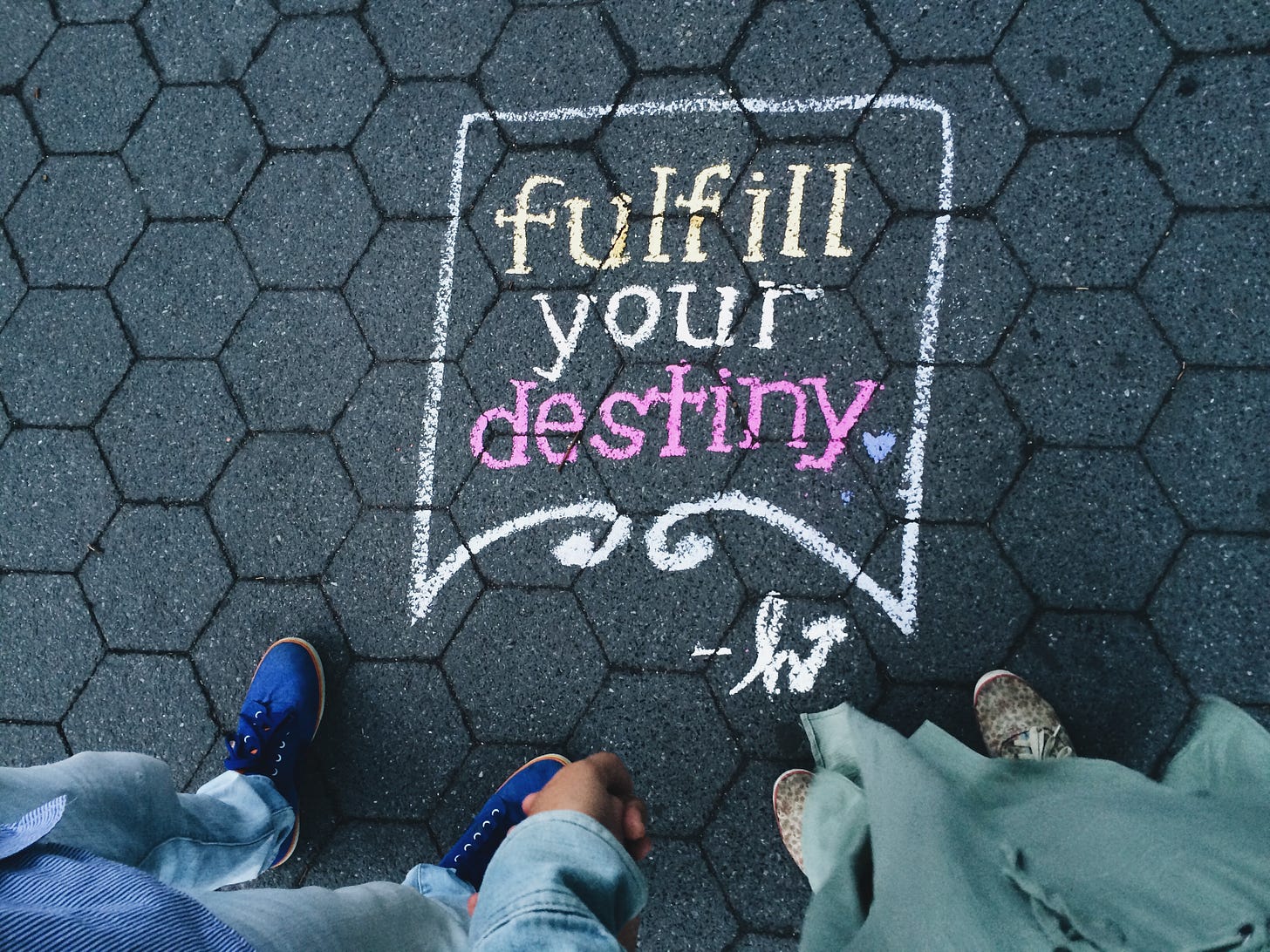 The words "fulfill your destiny" written in multicolored chalk on an asphalt surface. two pairs of shoes - one blue, one brown, stand in front of the drawing.