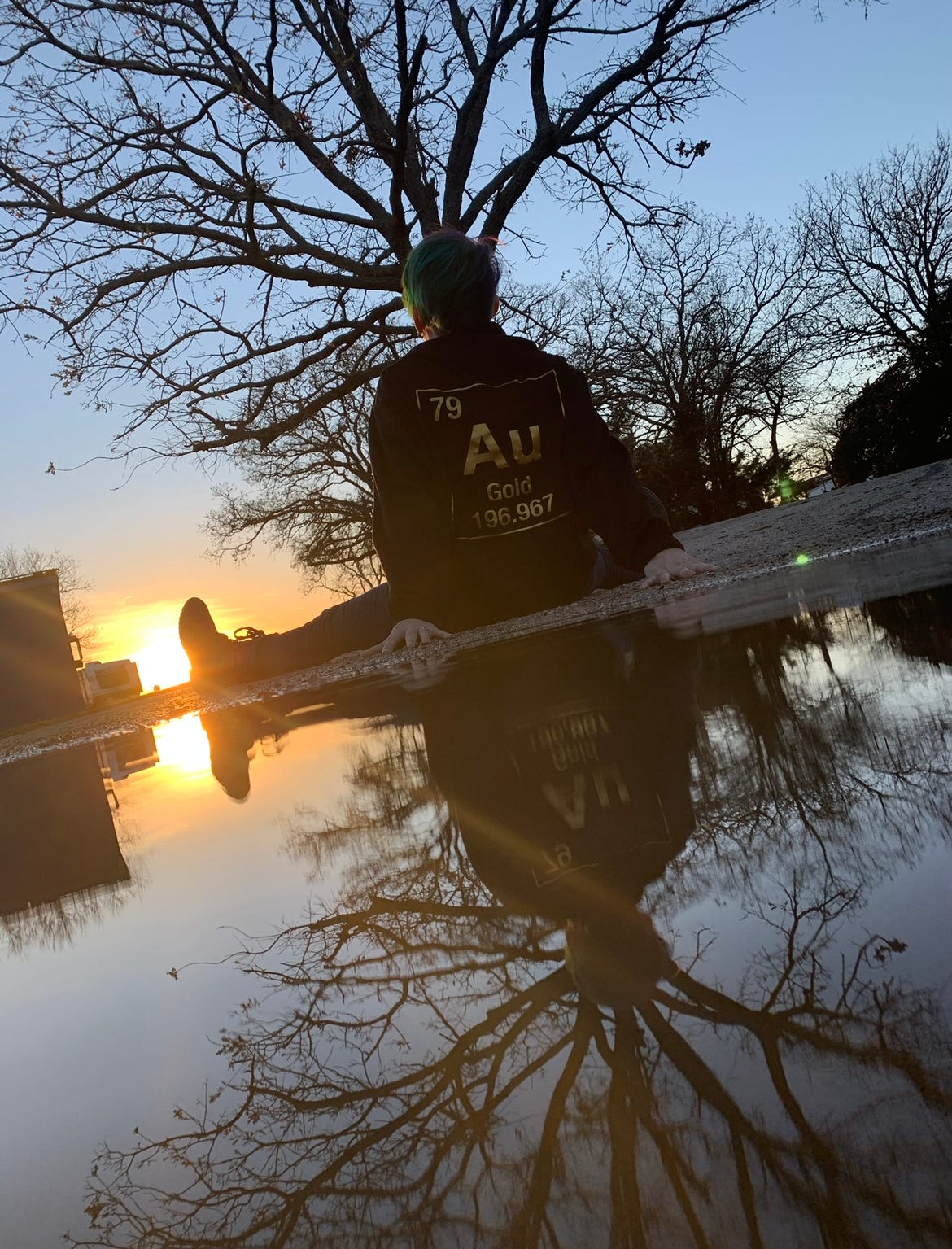 Lyric, sits watching a sunset, next to a puddle, reflecting back their green hair, and black sweater with an AU gold periodic table element in gold lettering on the back reflecting with bare trees in the puddle like a mirror