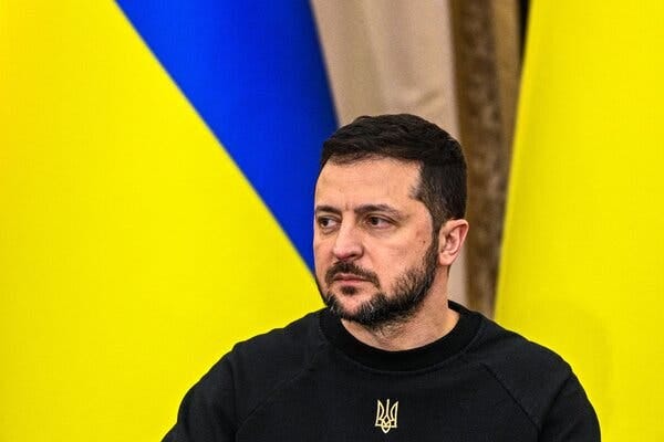 President Volodymyr Zelensky of Ukraine stands in front of two yellow and blue Ukrainian flags.