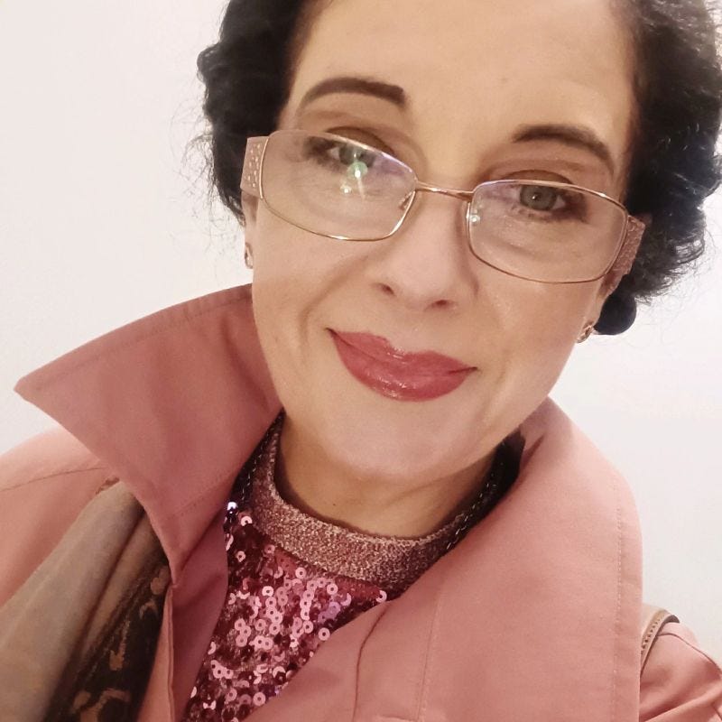 Woman with gold rim glasses on smiling and wearing pink coat and sparkly top