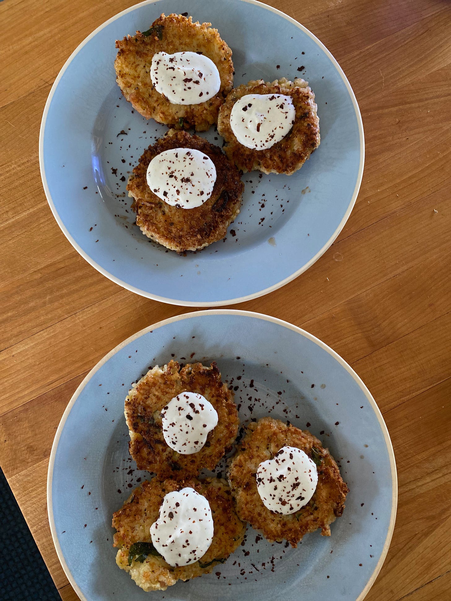 Two blue ceramic plates on a wooden counter, each holding three golden brown rice fritted topped with yogurt sauce and red pepper flakes.