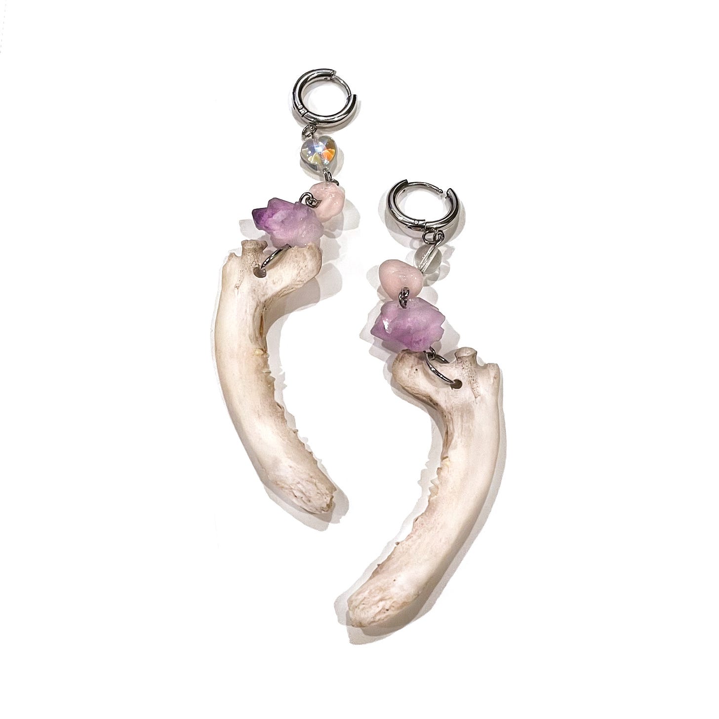 A pair of earrings made of raccoon jaws, Amethyst nuggets, rose quartz, glass beads, and silver toned huggie hoops resting on a white background.