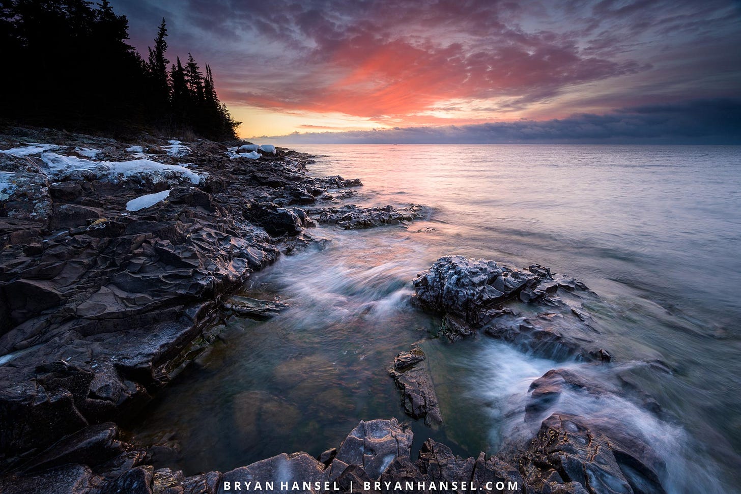 A photo of sunrise over a large body of water and a black basalt shoreline with a wave pool cut through its center. The sunrise is red and yellow.