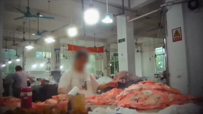 Garment worker in a Guangzhou factory from Zandland for Channel 4. The worker’s face is blurred and they stand in a factory in front a table piled high with clothing.