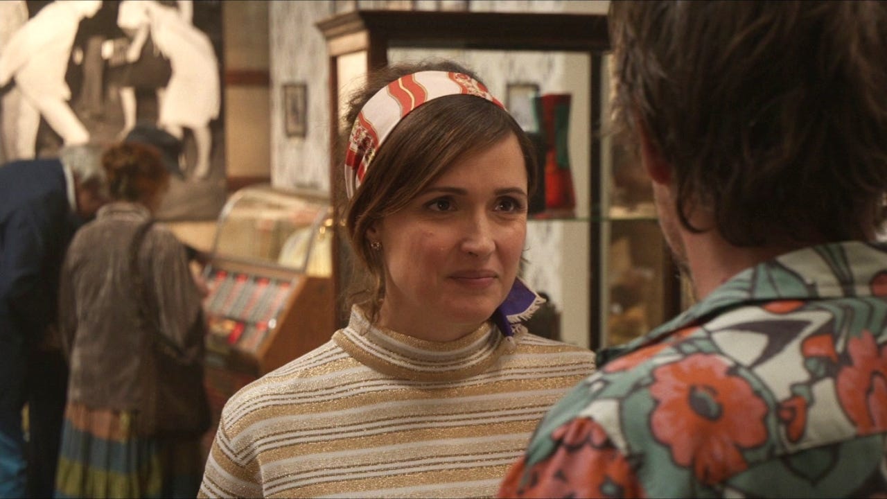Still from Juliet Naked where Annie is wearing her cute little striped glittery turtleneck and a scarf in her hair, very 1960s style, and Tucker is in a Hawaiian print shirt, and they're at the exhibit at her museum that she's been putting together on the Summer of '64.
