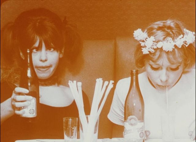 imagem em filtro sépia: duas adolescentes, uma de cabelo preto e outra loira usando coroa de flores, estão bebendo de garrafas. a de cabelo preto faz uma careta e a de coroa de flores bebe de um canudo