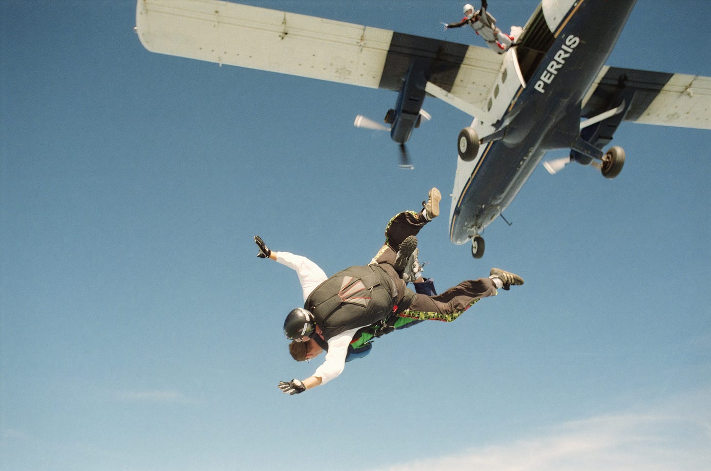 A photograph looking up at an aeroplane as people skydive out of it. A photograph looking up at an aeroplane as people skydive out of it.